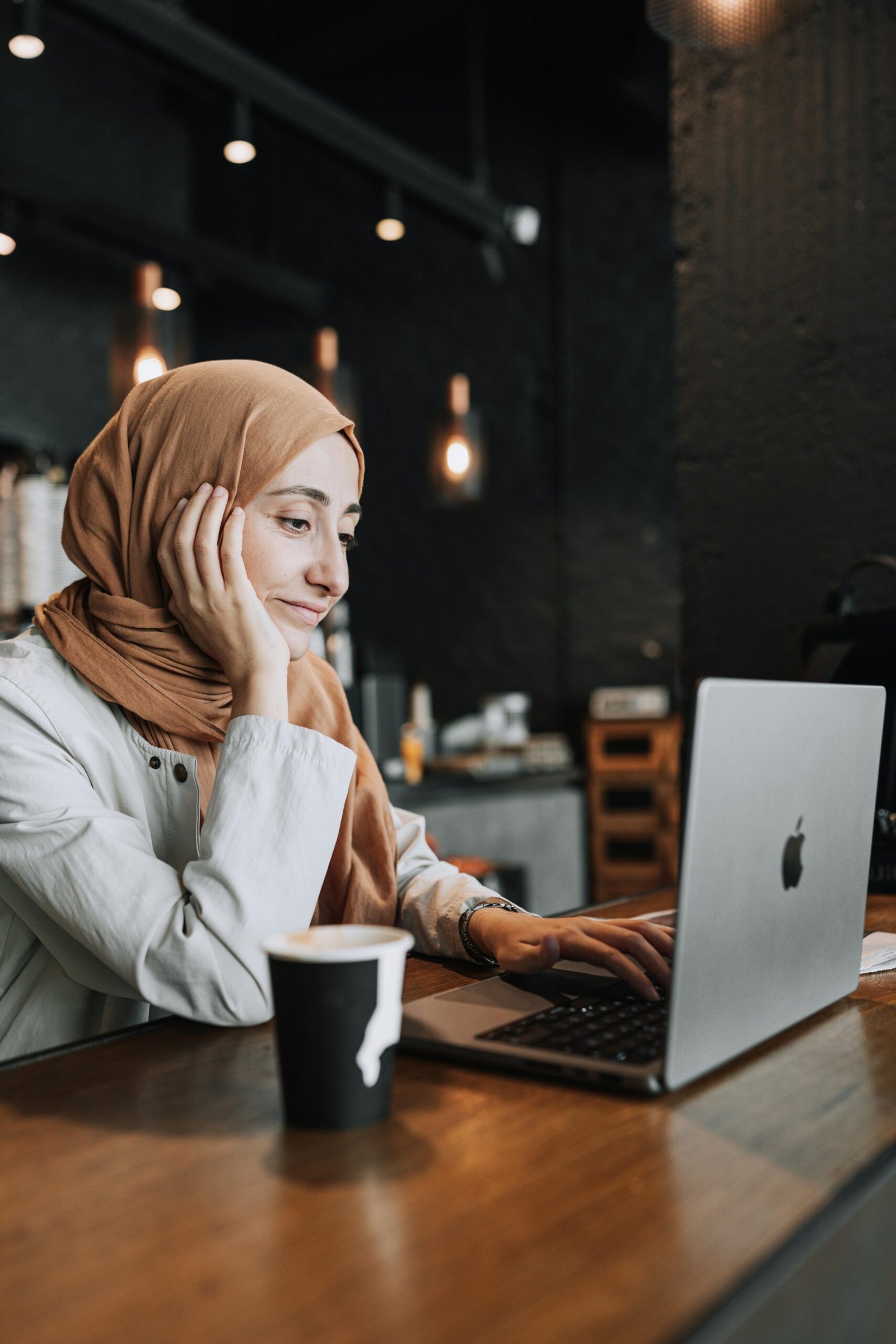 A woman in a headscarf works on her laptop in a warm café setting, enjoying a coffee.